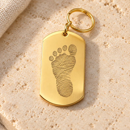 KI-Gestaltungsbeispiel: goldfarbener Edelstahl-Dogtag mit einem fiktiven Kinderfußabdruck als feine Line-Art-Gravur und Schlüsselring; kein gefertigtes Muster.