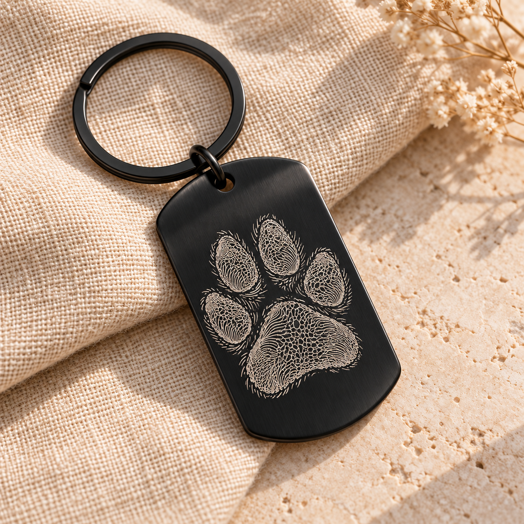 KI-Gestaltungsbeispiel: schwarzer Edelstahl-Dogtag mit einer fiktiven Pfote als feine Line-Art-Gravur und Schlüsselring; kein Foto eines gefertigten Musters.
