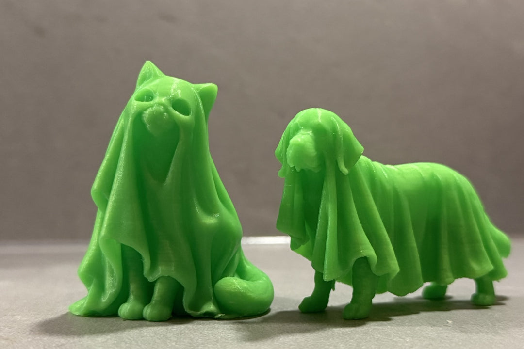 Originalfoto der Variante Grün nachleuchtend: sitzende Geisterkatze links und stehender Geisterhund rechts, beide mit ausgeformten Lakenfalten auf grauer Fläche. Weitere Farben: Blau nachleuchtend und Weiß ohne Leuchteffekt.